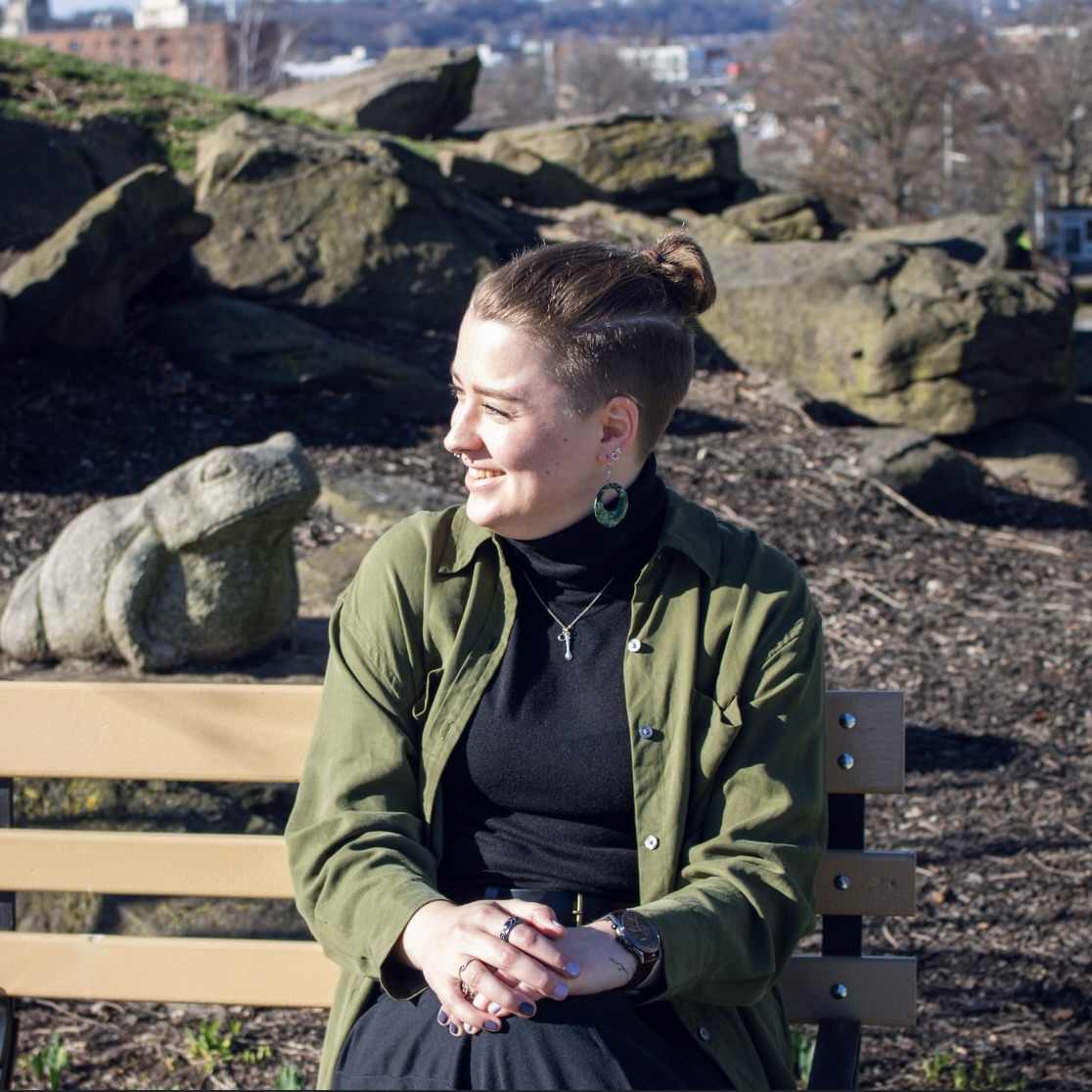 Lynn, with their light brown hair in a top knot and a fade around the sides. They have a septum piercing and are smiling widely, looking off to the left. They are wearing a green button down shirt, a necklace with a spoon charm, and green computer chip earrings.