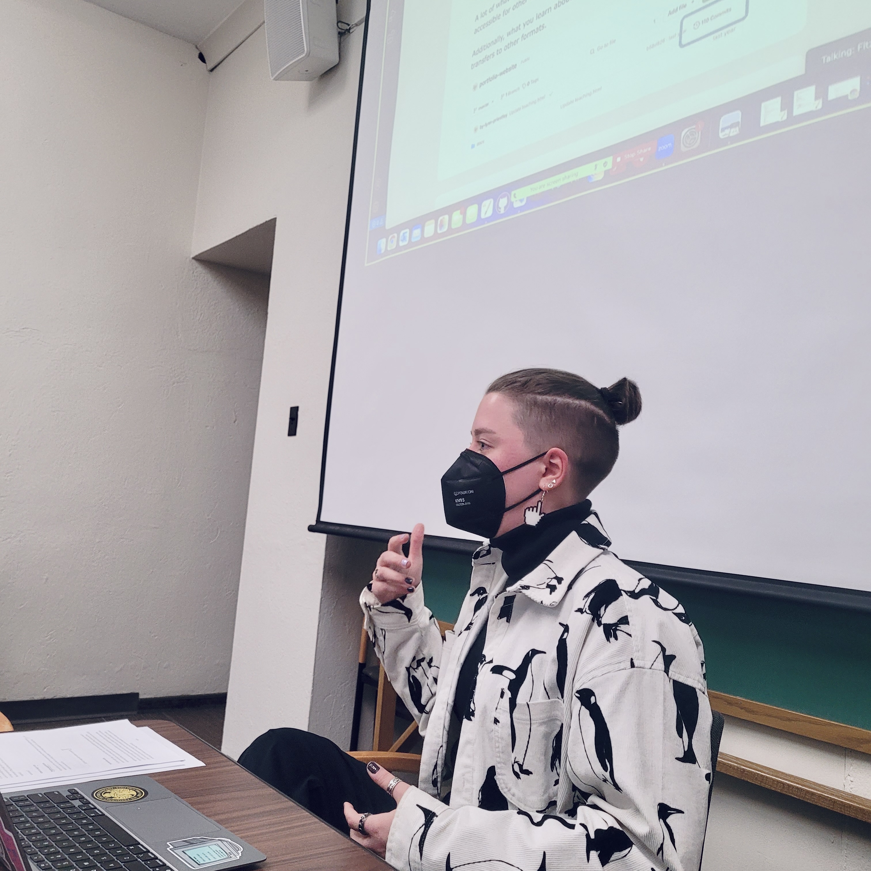 Lynn sitting and talking with their hands. Their hair is in a top knot with a fade around the sides. They are wearing a black n95 mask, big computer cursor earrings, and a black and white button down jacket with penguins on it. A screen with a demo document projected onto it is in the background.