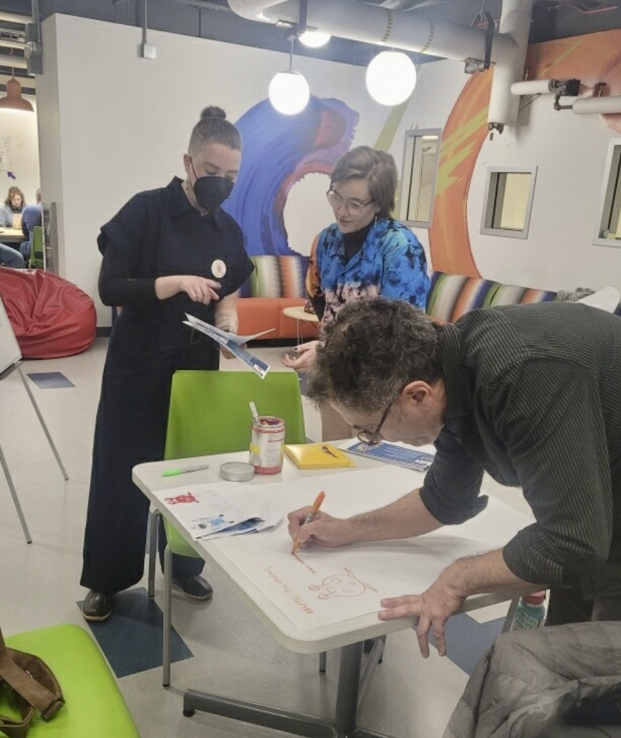 Lynn is standing with a DISCO participant, gesturing to a piece of paper in their hands that both individuals are looking at. In front of them, another DISCO designer leans over a large piece of paper, writing some notes next to a doodle of an alien persona.