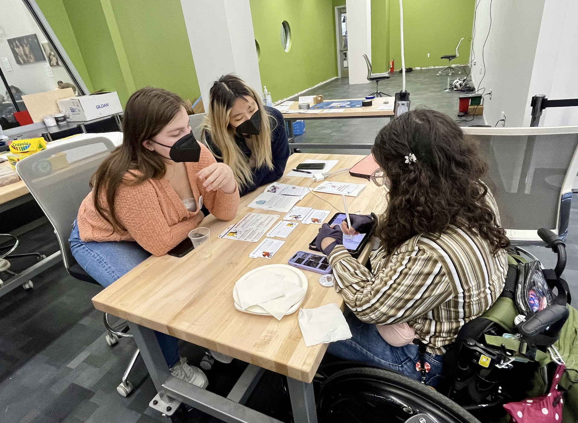 Three DISCO designers at a table with toolkit materials scattered about. One is drawing on an iPad, while the other two lean over to watch.