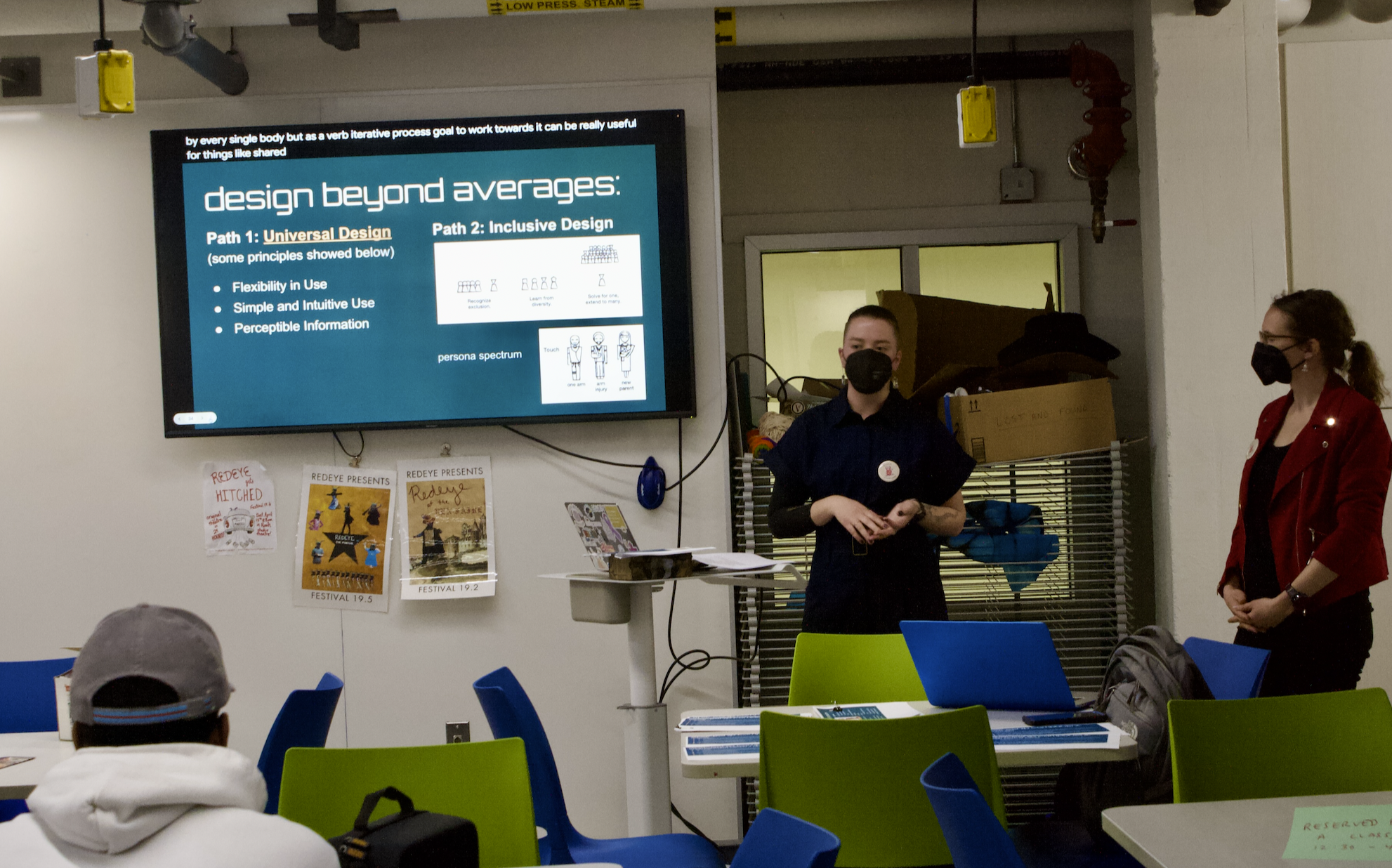 Lynn, a white person with brown hair in a top knot, and Dr. Fitz, a white woman with brown curly hair in a ponytail, stand to the right of a TV showing a slide titled 'Design Beyond Averages.' Both are wearing n95 masks, and Lynn is talking with their hands.