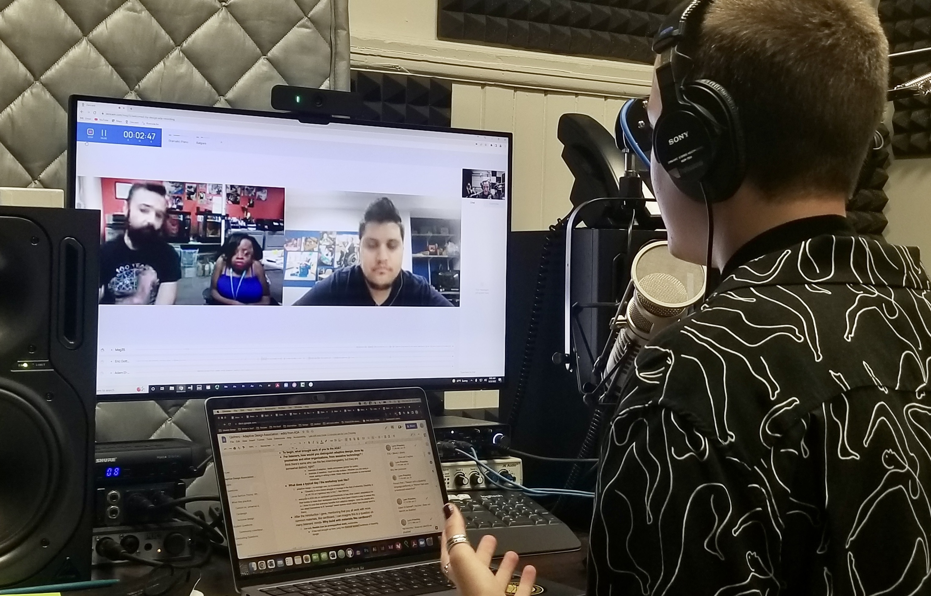 Eric, one of the team members of the Adaptive Design Association, is motioning with a hand, while the other two ADA members listen. All three are visible on remote video on the podcast studio monitor. Lynn is also motioning with their hand, and the back of their head is visible. A Google doc is pulled up on their laptop, with many comments on the side of it.