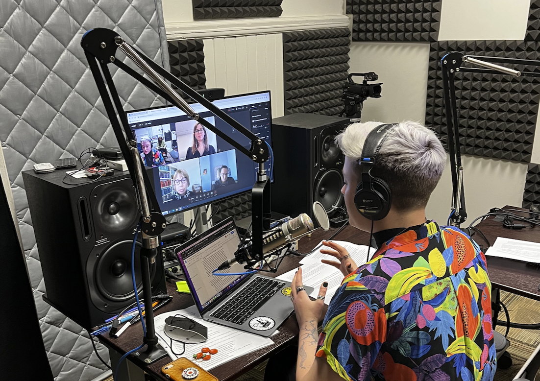 Lynn’s eyes are looking down at the interview notes in front of them, as they talk into the mic while also gesturing with their hands. The microphone slightly obstructs the view of Meryl Alper, Bess Williamson, and Elizabeth Guffey, the roundtable on the monitor.