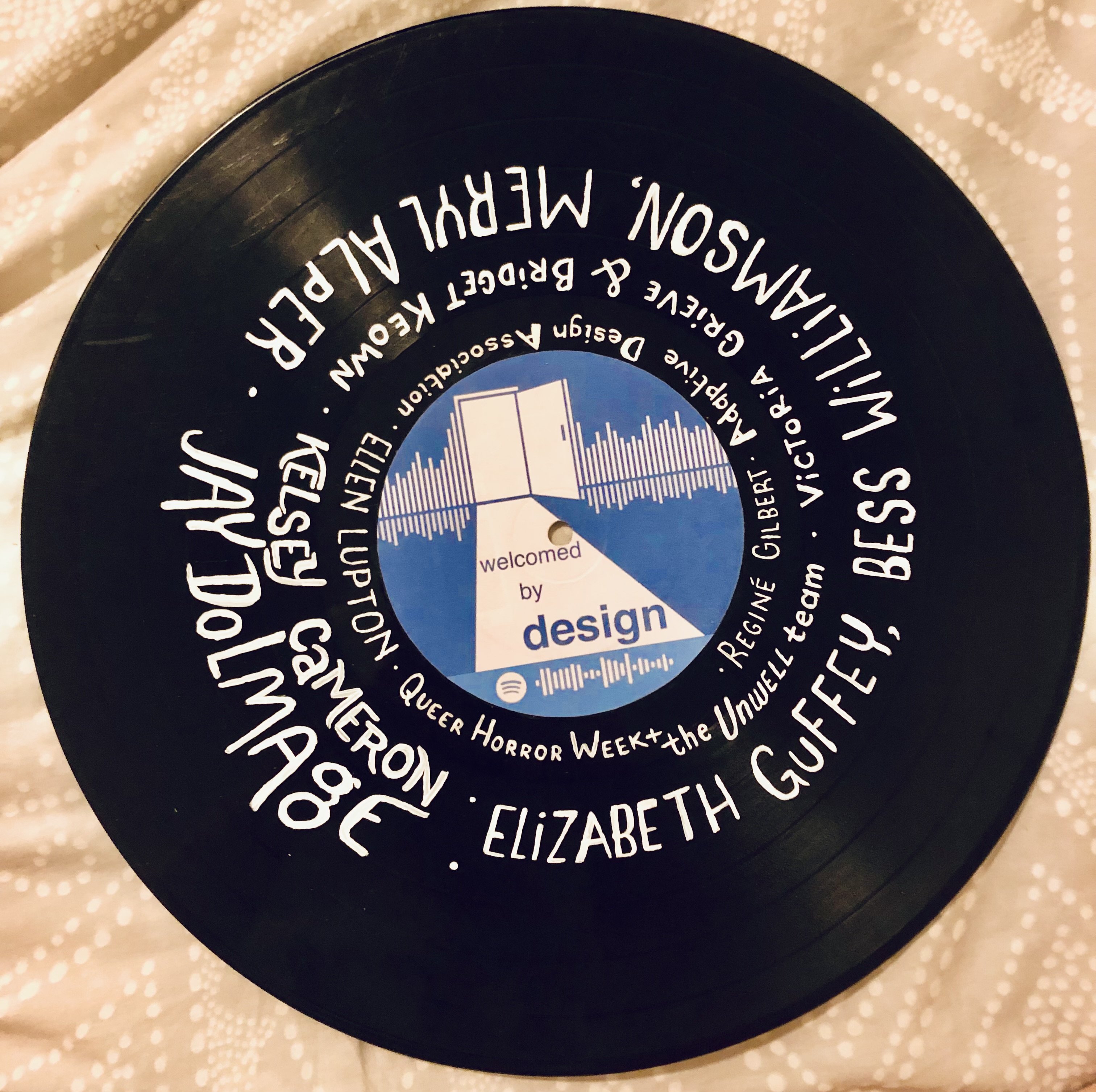 Vinyl with the Welcomed by Design cover art as its label. The names of podcast guests are painted around the vinyl: Reginé Gilbert, Adaptive Design Association, Ellen Lupton, Queer Horror Week + the Unwell team, Victoria Grieve and Bridget Keown, Kelsey Cameron, Elizabeth Guffey, Bess Williamson, and Meryl Alper, and Jay Dolmage